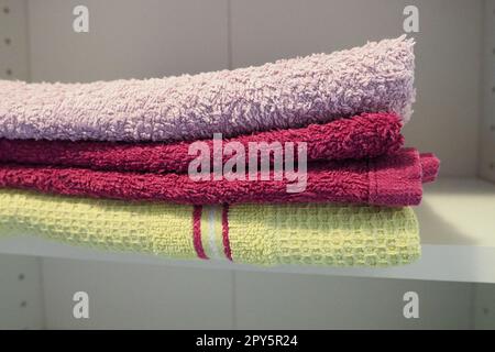 Towels on a shelf in a white cabinet. Clean ironed pink and yellow towels folded in a pile. Organization of household items in the bathroom or closet. The result of the work of a housewife. Stock Photo