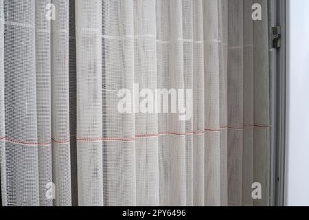 White coarse cotton or linen curtains made of weaves of thin threads. Simple compact economical apartment interior. Window decoration in the room. Red stripe. Stock Photo