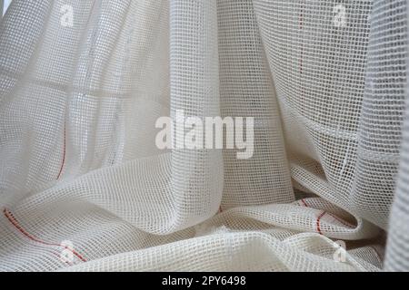 White coarse cotton or linen curtains made of weaves of thin threads. Simple compact economical apartment interior. Window decoration in the room. Red stripe. Stock Photo