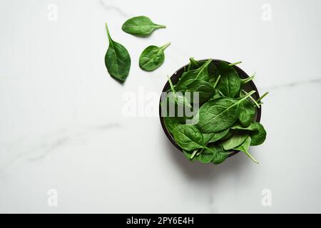 A bunch of Fresh baby spinach leaves in the in a black ceramic bowl on white marble background. Top view and flat lay photo with copy space Stock Photo