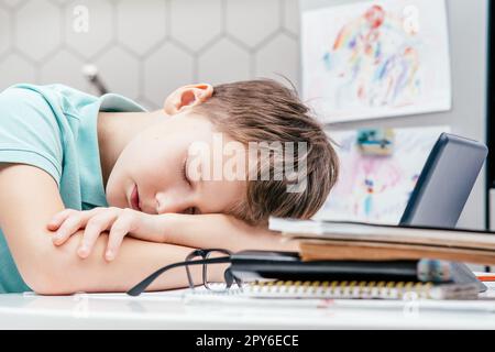 Side view of young preteen exhausted boy pupil sleeping on folded hands on desk near notebooks, glasses, tablet at home. Stock Photo