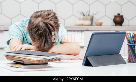 Portrait of young preteen tired boy pupil lying sleeping on folded hands on desk near notebooks, glasses tablet at home. Stock Photo