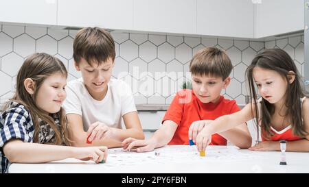 Funny little school friends play board game with dice and chips at kitchen table. Children throw dice and move figure. Stock Photo