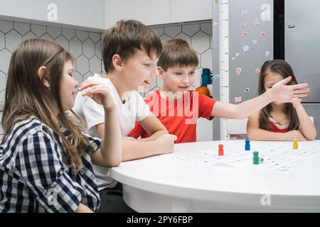 Happy little school friends play board game on paper map with dice and chips at table, side view. Interesting table game Stock Photo