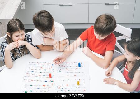 Friends play board game with dice and chips on table, from above view. Kids look at boy throw dice and move figure. Stock Photo