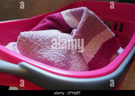 Towels in the laundry basket. Blue and pink cotton terry towels are thrown into a pink plastic basket. Housekeeping. Storing and separating laundry before washing. Light from above from open window. Stock Photo