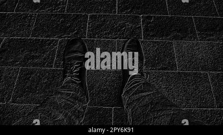 Monochrome black and white image. Feet in boots against the background of street tiles or the road. Two legs of a seated man, photographed from above while sitting. Stock Photo