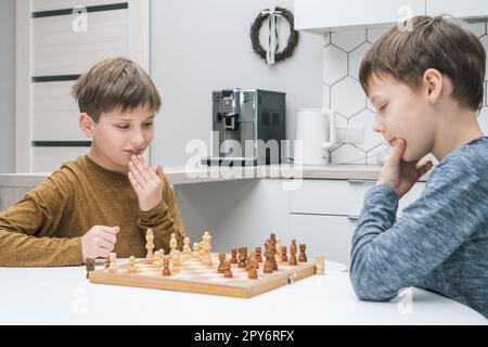 Thinking schoolboys play chess sitting at kitchen table, side view. Wooden chessboard with black and white figures. Stock Photo