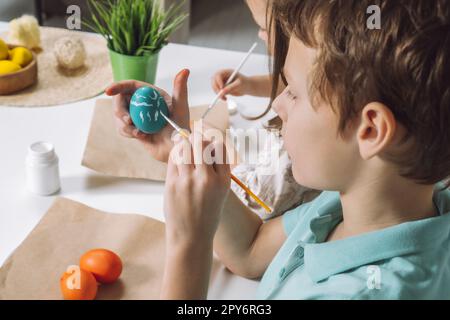Cropped portrait of boy hand, painting multi coloring Easter hens eggs, brown paper plate table, paintbrush Stock Photo