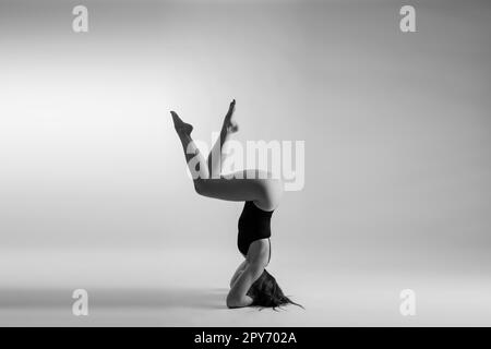 Young slim female in black bodysuit makes yoga stretching exercise on white studio wall background. Stock Photo