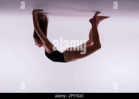 Young slim female in black bodysuit makes yoga stretching exercise on white studio wall background. Stock Photo