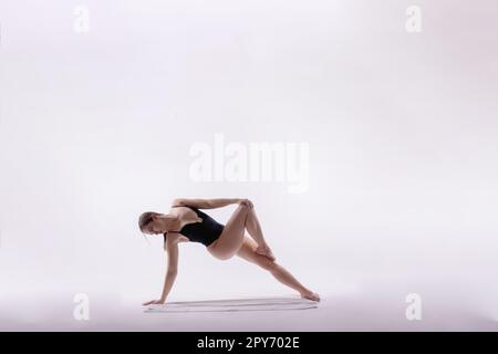Young slim female in black bodysuit makes yoga stretching exercise on white studio wall background. Stock Photo
