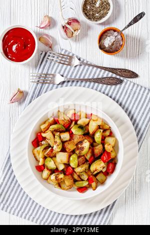 Pan-Browned Potatoes with bell red pepper and whole garlic cloves in white bowl on white wooden table, vertical view from above Stock Photo