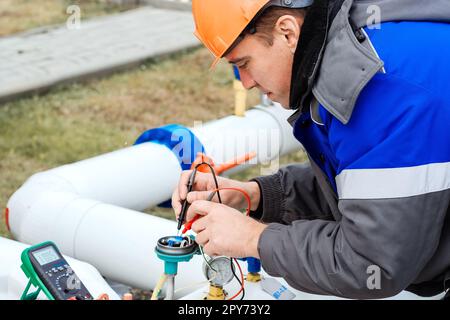 Caucasian electrician in helmet measures voltage with multimeter on sensors of gas distribution station outside. Genuine workflow Stock Photo