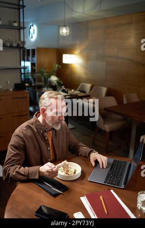 Mature man using laptop while having lunch sitting at table in restaurant Stock Photo