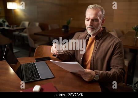 Mature man drinking water and reading contract while working on laptop at table in cafe Stock Photo