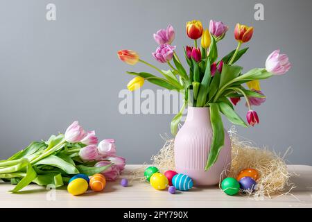 A bunch of tulips in a vase standing on the table and colourful eggs lying on it. Stock Photo