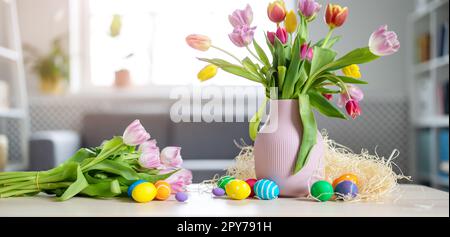 A bunch of tulips in a vase standing on the table and colourful eggs lying on it. Stock Photo