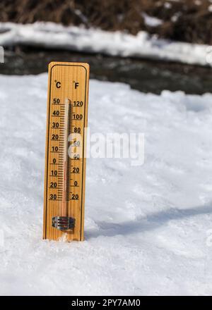 Wooden thermometer standing in ice on partially frozen river, sun shining, showing +3 degrees. Image to illustrate winter leaving, snow slowly thawing, warmer days coming Stock Photo