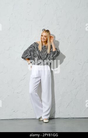 Cheerful and positive woman in striped blouse and wide trousers leaning against textured wall Stock Photo