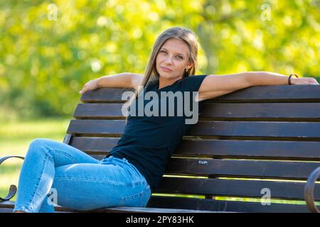 A girl sits on a bench in a sunny park and looks into the frame Stock Photo