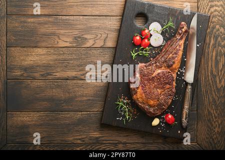 Tomahawk steak. Sliced grilled tomahawk beef steak with baked cherry tomatoes, herbs and salt on old wooden background. Preparing to grill.  Top view Stock Photo