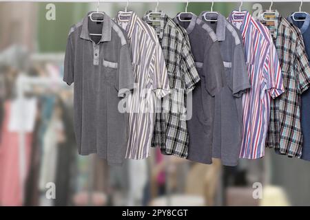 Clothes on rack. Clothing rack with various colorful male casual summer shirts over abstract blurred clothes shop. All hanging neatly on hangers at cloth rail. Space. Shopping. Stock Photo