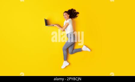 Cheerful Young Woman Using Laptop Computer Jumping Over Yellow Background Stock Photo