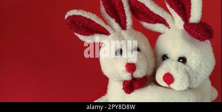 Two white plush rabbits cuddling. Dark red background. Children's toys are white. Greeting card on Valentine's Day. Mutual love and affection. Declaration of love Stock Photo