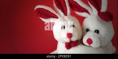 Two white plush rabbits cuddling. Dark red background. Children's toys are white. Greeting card on Valentine's Day. Mutual love and affection. Declaration of love. Easy vignetting Stock Photo
