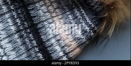 Knitted hat with a pumpkin close-up on a gray main background. Winter clothing advertising. Winter season. White gray, beige, brown and black tones. Big loops. The bean-brown pumpon from natural fur Stock Photo