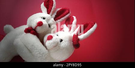 Two white plush rabbits cuddling. Dark red background. Children's toys are white. Greeting card on Valentine's Day. Mutual love and affection. Declaration of love. Easy dark vignetting Stock Photo