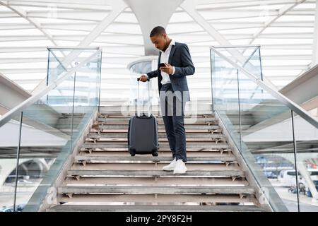 Young Black Businessman With Smartphone Walking Down Stairs At Airport Terminal Stock Photo