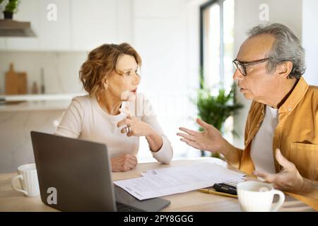 Worried senior spouses checking financial documents with laptop, sitting in kitchen, suffering problems Stock Photo