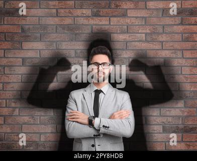 Businessman and shadow of strong muscular man behind him on brick wall. Concept of inner strength Stock Photo