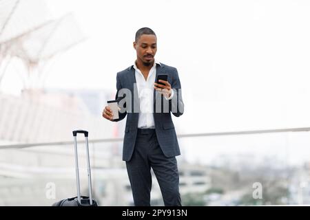 Young Black Businessman Using Smartphone And Having Coffee While Waiting At Airport Stock Photo