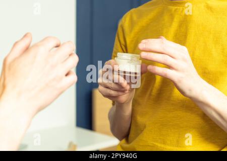 Hands of middle aged man holding jar of moisturizing cream. Crop male portrait in room mirror. Facial care concept. Stock Photo