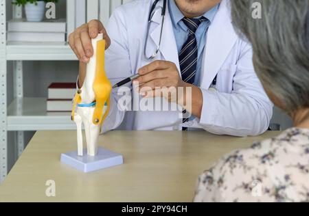 Orthopedic Surgeon in white gown and stethoscope pointing at anterior cruciate ligament on deluxe functional knee joint model, present to the elderly patient about treatment of the knee with surgery. Stock Photo