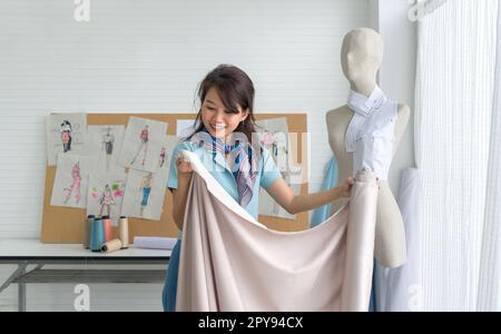 Young asian dressmaker in striped scarf check the quality of beige fine fabric from the roll. Fashion designer room. Stock Photo