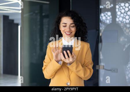 Close up of successful smiling hispanic woman using phone, businesswoman at workplace holding smartphone reading news online standing near window in yellow jacket. Stock Photo
