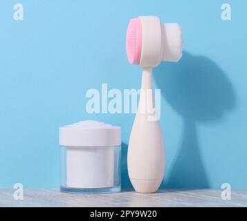 A white jar of cream and a facial cleansing brush on a blue background, items for cosmetic procedures. Stock Photo