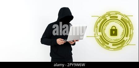 Male programmer in black hoodie stands and holds laptop. Hacker with hood on his head typing text on computer. Futuristic digital design on the wall. Stock Photo