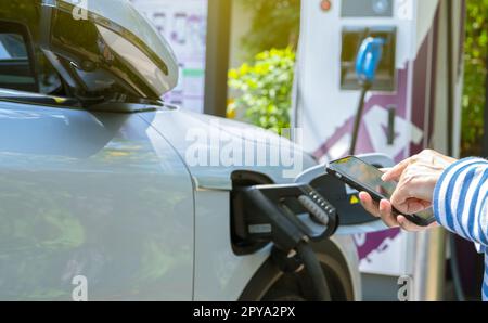 Woman using smartphone paying service in app on blur EV car charging at electric car charging station. EV car charging point. Electric vehicle fast charge. Sustainable power for reduce carbon emission Stock Photo