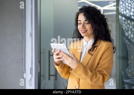 Young beautiful hispanic businesswoman standing in the office in the corridor. He holds a tablet in his hands, uses it, types, smiles Stock Photo