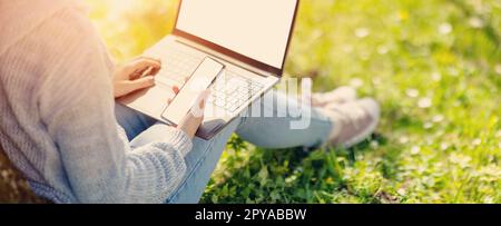 Woman with laptop and smartphone sitting on the grass in the natural park. Stock Photo