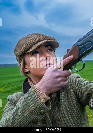 Portrait of a thirty plus year old woman with a shotgun stood in a field against a stormy blue sky Stock Photo