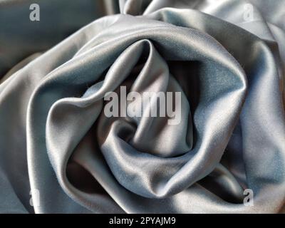 Silky to the touch fabric of gray - blue color with a brilliant shimmer. Natural lighting, light and shadow. The material is casually folded into a circle, resembling a rose bud. Polyester Textile. Stock Photo