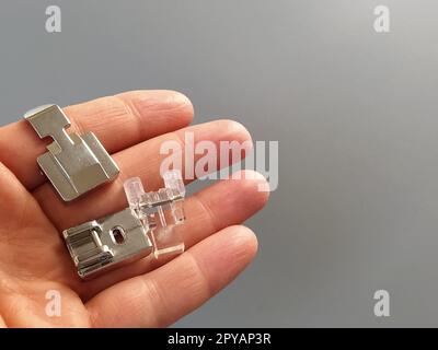 A set of feet for an electric sewing machine in a female hand. Gray backdrop. Standard sewing or zigzag foot, for sewing in zippers, overlock foot and for buttons Stock Photo