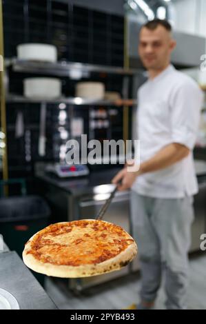 Young male chef holding freshly baked pizza on long shovel selective focus Stock Photo