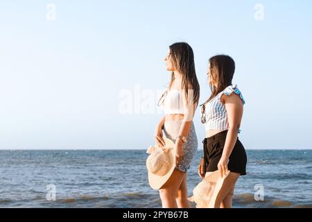 Two young female friends standing side by side on a sunny beach, smiling and looking out to sea Stock Photo